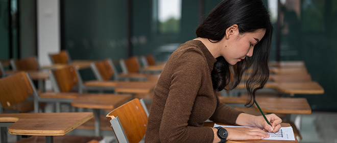 A female in a brown shirt writing an exam while sitting at a desk in an exam room with desks in the background.